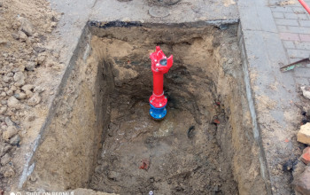 Zdjęcie przedstawia duży, kwadratowy wykop w ziemi, otoczony luźną glebą i fragmentami chodnika. W centralnej części wykopu zainstalowany jest nowy, czerwony hydrant z niebieską podstawą.
