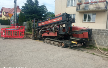 To zdjęcie przedstawia czerwono-czarną maszynę gąsienicową, która jest wiertnicą horyzontalną do wykonywania przewiertów sterowanych pod ziemią. Maszyna stoi na utwardzonym poboczu. 
