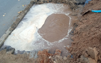 Na zdjęciu widoczny jest duży, prostokątny wykop pełen brudnej wody i błota, znajdujący się przy krawężniku drogi asfaltowej.
Powyżej wykopu widoczny jest przód żółtej koparki.