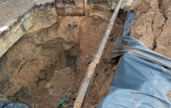 To zdjęcie przedstawia głęboki wykop ziemny w pobliżu drogi, wypełniony brudną ziemią i błotem. W górnej części obrazu widoczna jest żółta koparka.