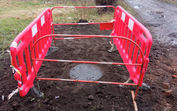 Czerwone, plastikowe barierki ochronne otaczają okrągłą, metalową pokrywę uszkodzonej studni rewizyjnej na kanalizacji sanitarnej. Teren wokół jest rozkopany i zabłocony.