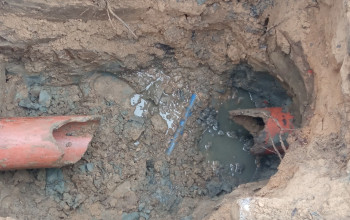 Dwa popękane, pomarańczowe przewody kanalizacji deszczowej są częściowo zakopane w błotnistym wykopie ziemnym.
W miejscu uszkodzenia wokół rur zalega woda i luźna ziemia.