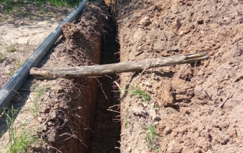 Wąski i głęboki wykop ziemny biegnie przez plac budowy w kierunku żółtej koparki znajdującej się w tle.
Obok wykopu ułożone są długie, szare rury, a nad nim leży drewniany pal.