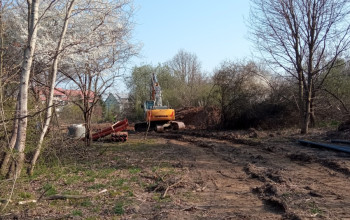 Duża, żółta koparka gąsienicowa stoi na błotnistym terenie, gdzie prowadzone są prace przy budowie sieci wod-kan.
Na placu widać ślady gąsienic, fragmenty rur.
